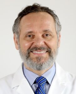 A doctor in a blue shirt, blue tie and white coat with a grey beard smiling